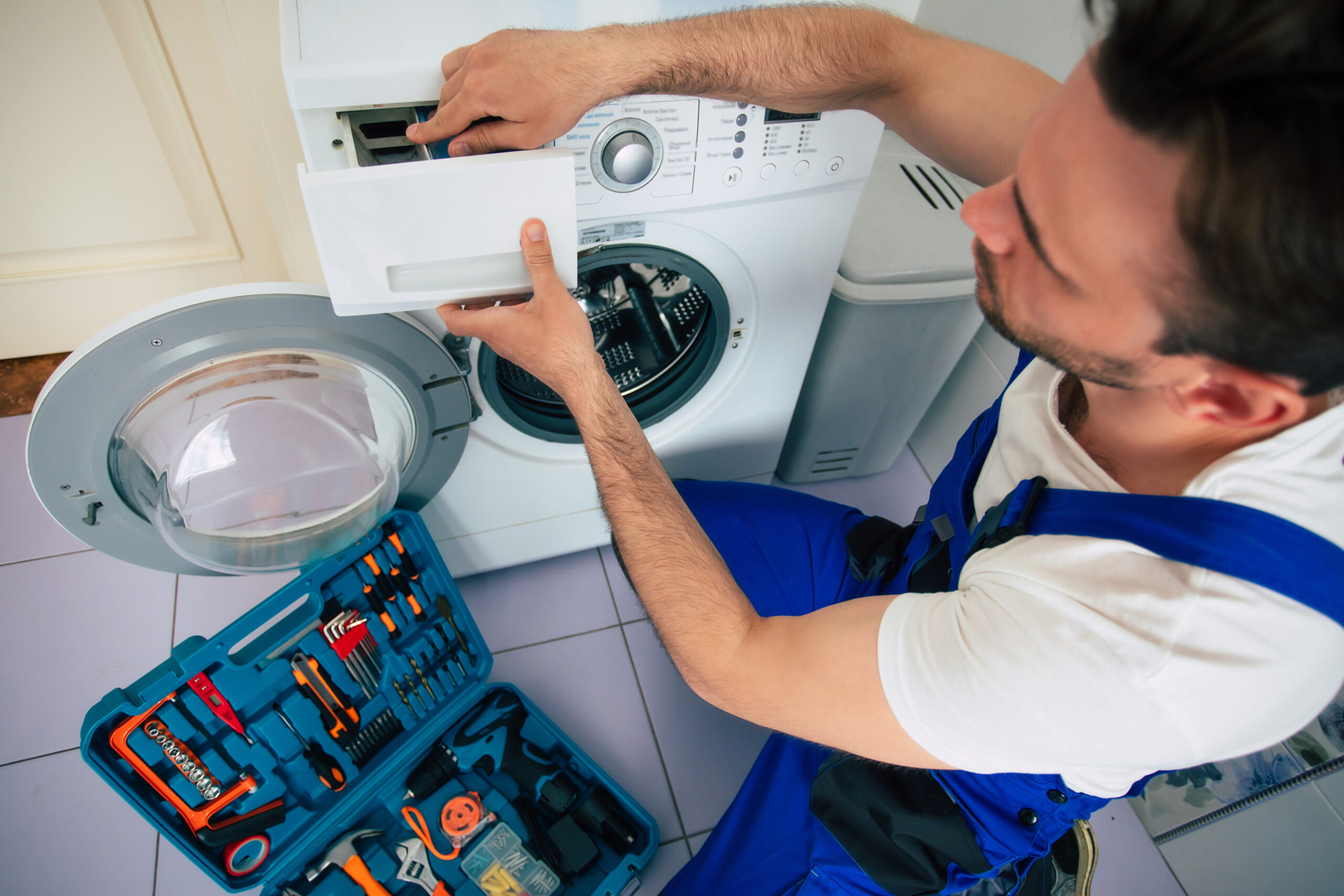The young handsome repairman in worker suit with the professional tools box is fixing the washing machine in the bathroom