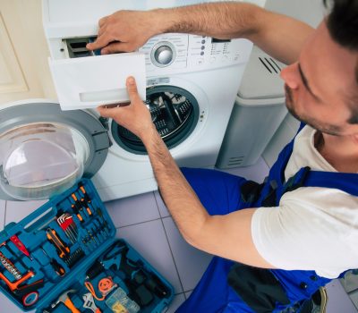 The young handsome repairman in worker suit with the professiona The young handsome repairman in worker suit with the professional tools box is fixing the washing machine in the bathroom